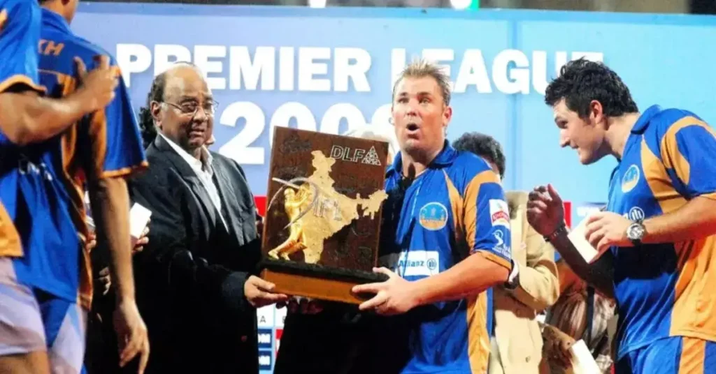 An iconic photo of Shane Warne lifting the IPL 2008 trophy with the Rajasthan Royals team celebrating around him. ALT: Shane Warne lifting IPL 2008 trophy